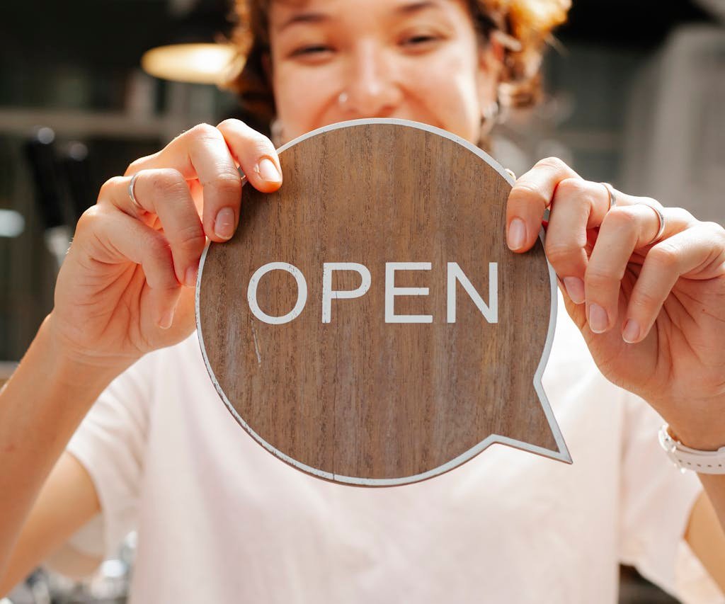 Happy woman holding an 'Open' sign at a café, inviting customers indoors.